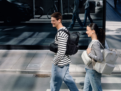 Zwei lächelnde Personen in gestreiften Shirts überqueren eine Straße. Der Mann trägt einen schwarzen Rucksack von Kapten & Son, die Frau einen hellgrauen Rucksack von Kapten & Son.