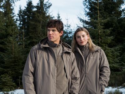 Ein Mann und eine Frau stehen in einer verschneiten Waldlandschaft und tragen passende taupefarbene Outdoorjacken von Kapten & Son.