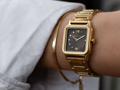 Nahaufnahme eines Handgelenks mit einer rechteckigen Uhr von Kapten & Son in Gold mit braunem Zifferblatt und römischen Ziffern, kombiniert mit einem goldenen Armreif.